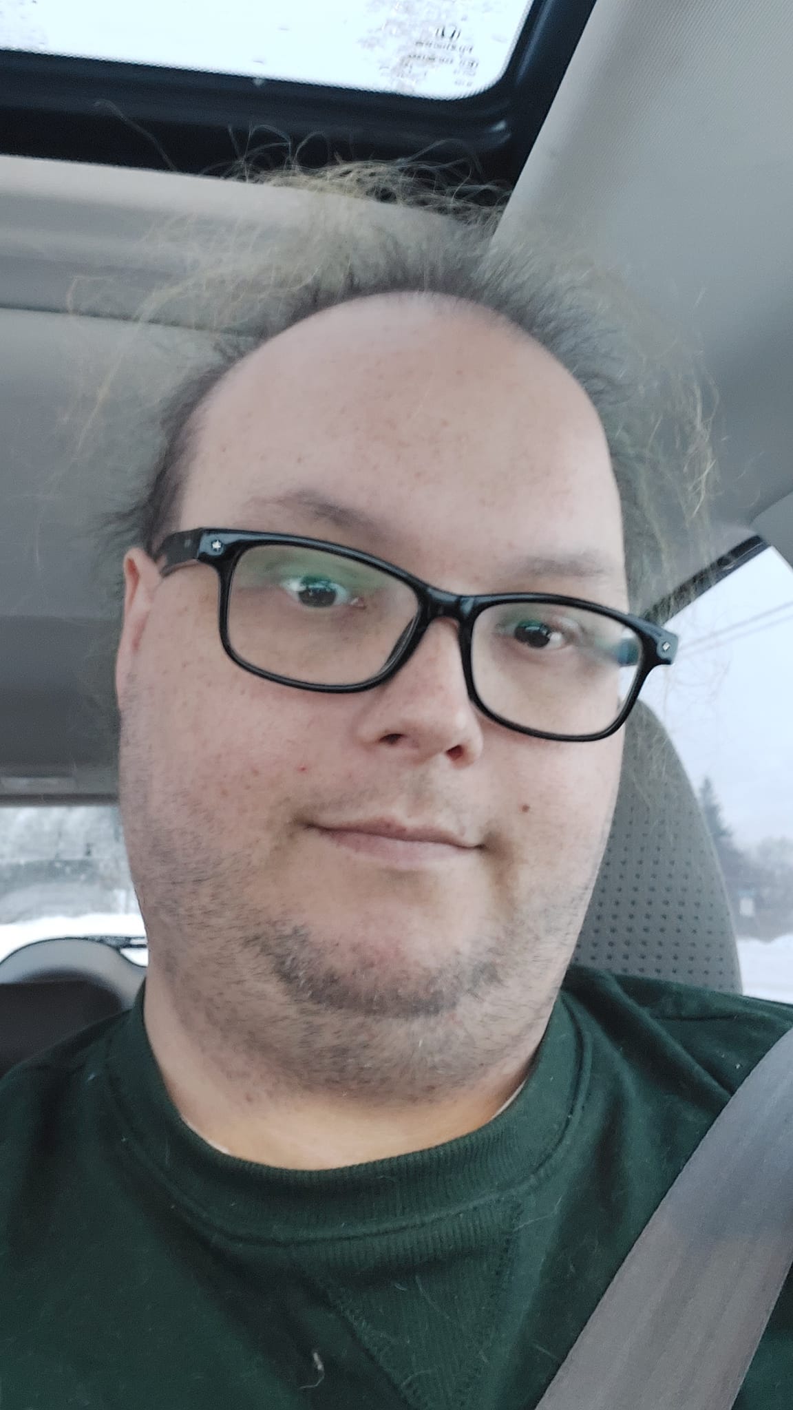 A man with thin hair is wearing a green sweater and sitting in a car, it is clearly winter outside